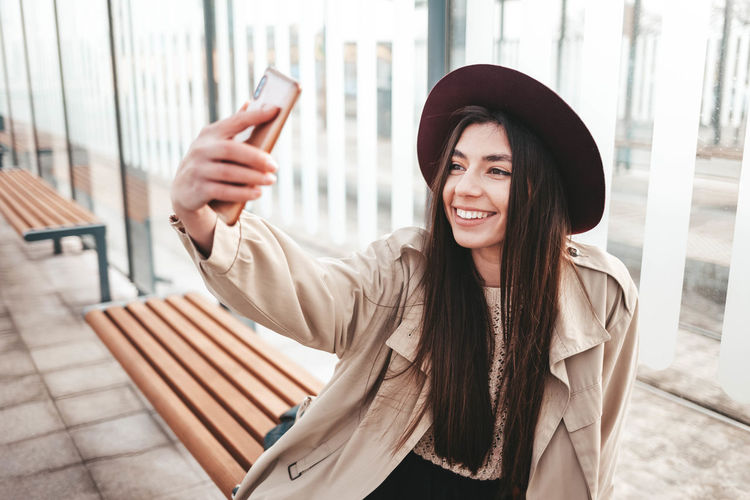 Stylish girl in hat makes selfie while sitting at a public bus stop