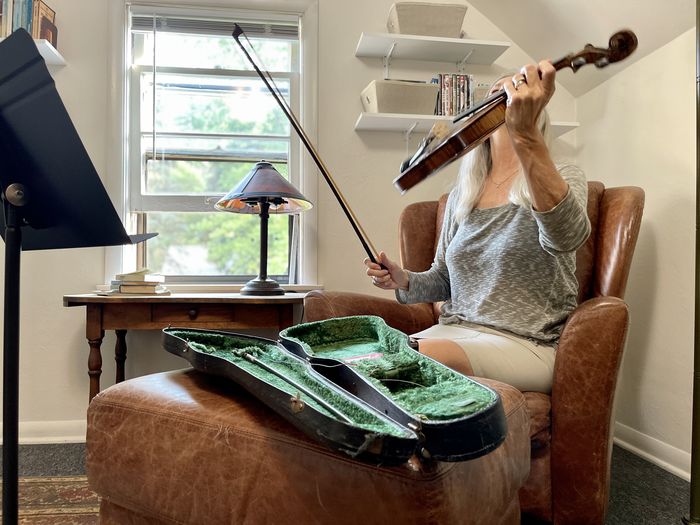 Active senior woman picks up violin to play indoors with music stand, bow and open violin case.
