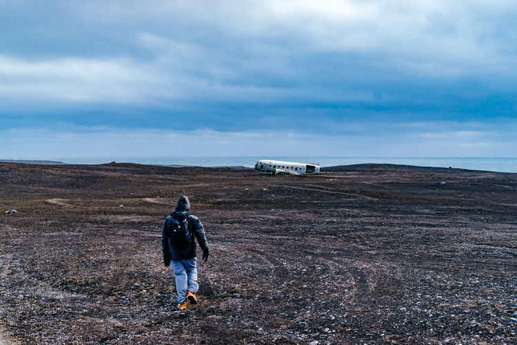 Full length rear view of man walking towards abandoned military airplane at beach against cloudy sky during winter