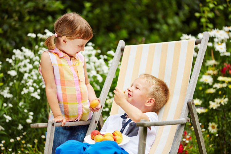 Girl with brother eating fruit while sitting on deck chair at park