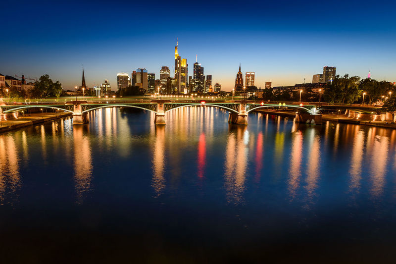 Illuminated bridge over river by buildings against sky in city