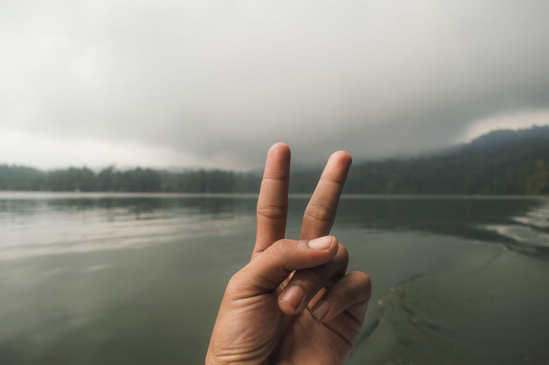 Cropped hand gesturing peace sign by lake against cloudy sky