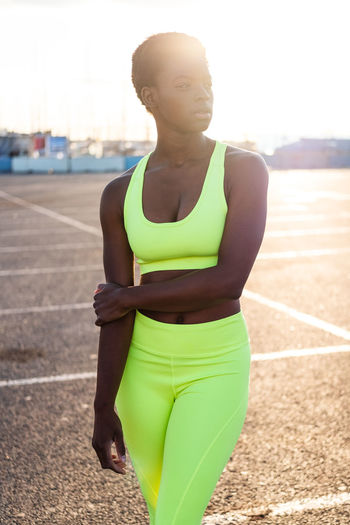 Serious focused african american woman in vibrant yellow sportswear looking away and contemplating in sunbeams alone on street against urban environment