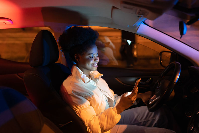 Smiling happy african american girl using smartphone, sharing news in social media while driving car