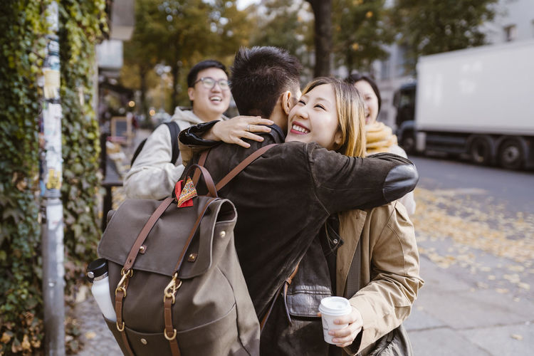 Happy young woman embracing male friend wearing backpack while standing at sidewalk