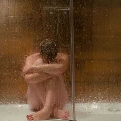 Full length of shirtless depressed woman sitting in bathroom seen through wet glass window