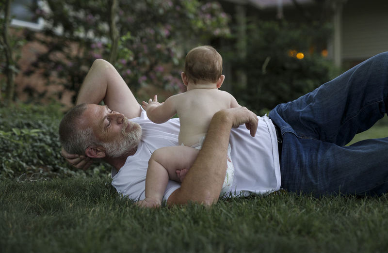 Grand father playing with baby boy while lying on grass in backyard
