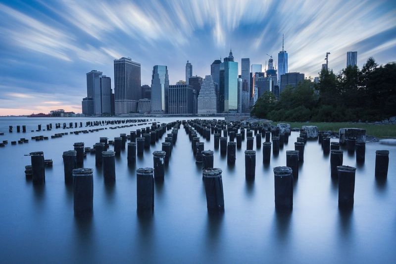 Wooden posts in sea by city skyline against sky at dusk