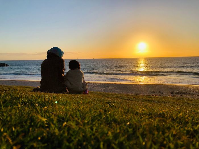Rear view of woman sitting with daughter at beach during sunset