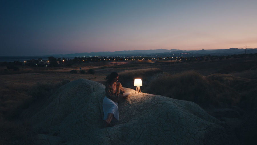 Woman reads a book in the evening on a rock with lamp