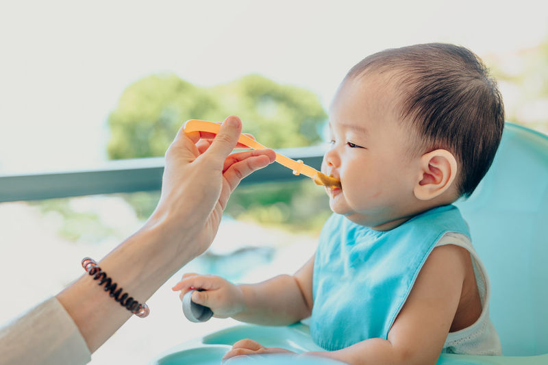 Cropped hand of mother feeding baby son sitting on high-chair at home