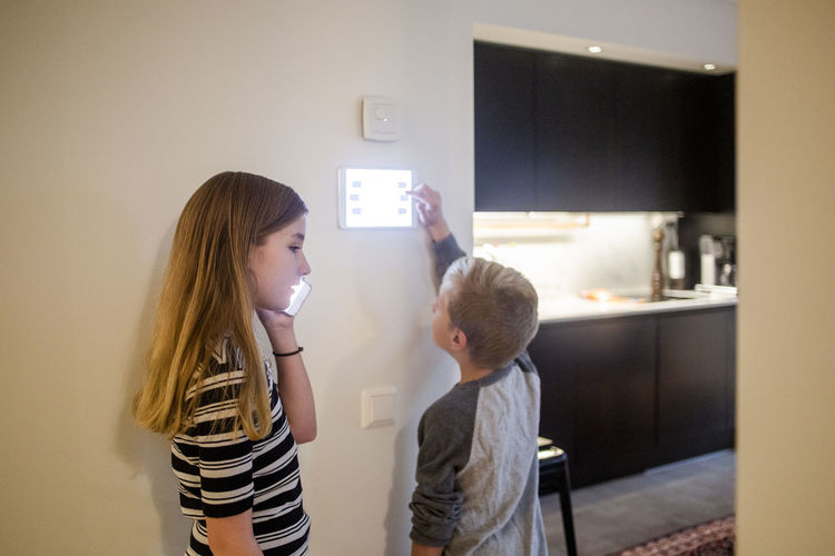 Girl talking on smart phone while brother using digital tablet on wall at home