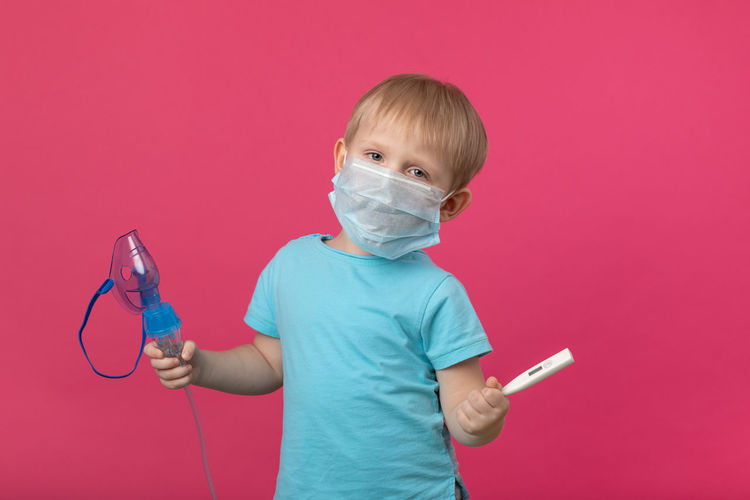 A blond child with an inhaler and a thermometer in his hand on a plain pink background. studio. 
