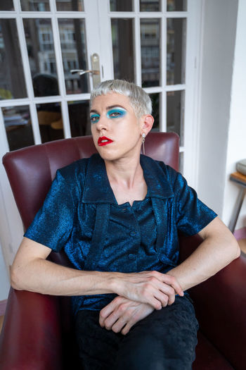 Portrait of confident androgynous man wearing makeup sitting on armchair at home