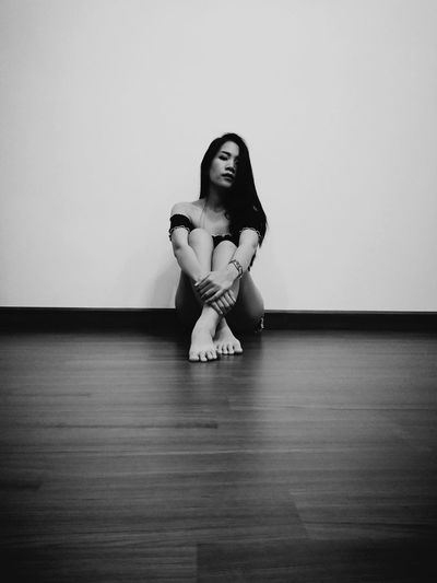 Full length portrait of young woman sitting on hardwood floor against white wall at home