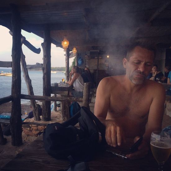 Shirtless man sitting by drink in glass and sea