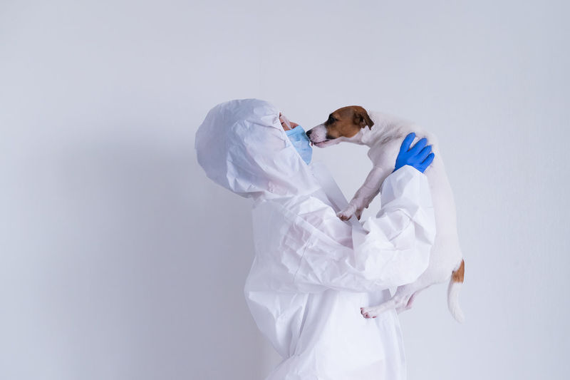 Woman in biohazard suit kissing dog against white background