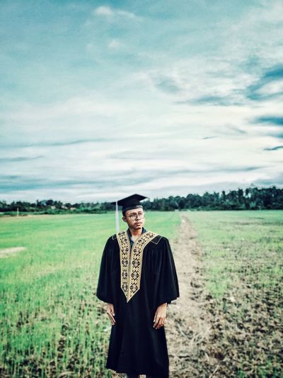 Young man wearing graduation gown while standing on grassy field against sky