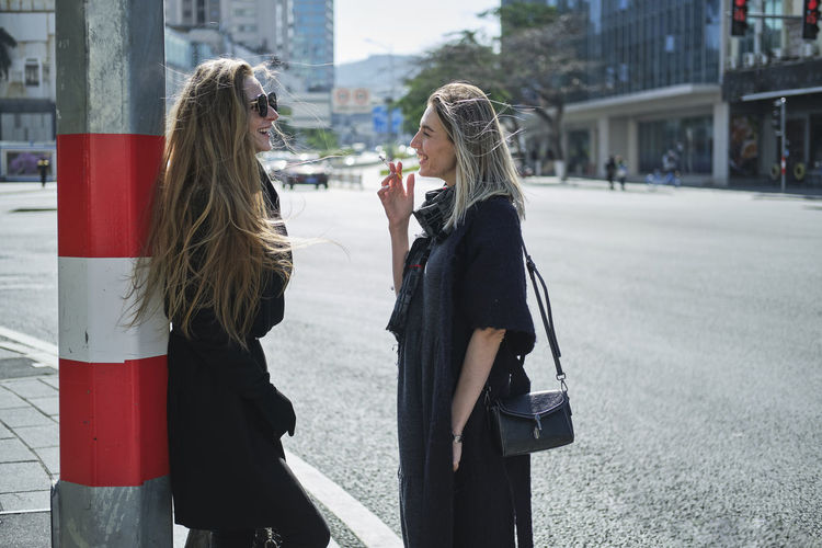 Side view of young content stylish woman with cigarette speaking with best female friend while looking at each other on street