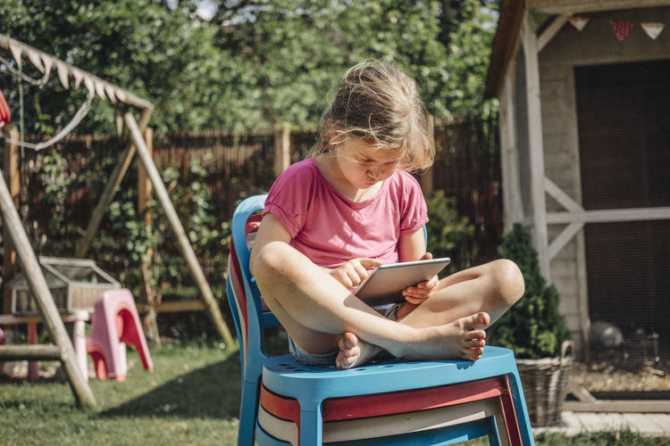 Girl sitting on stack of chairs in garden using tablet