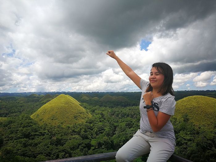 Smiling young woman with hand raised sitting on railing against cloudy sky at chocolate hills