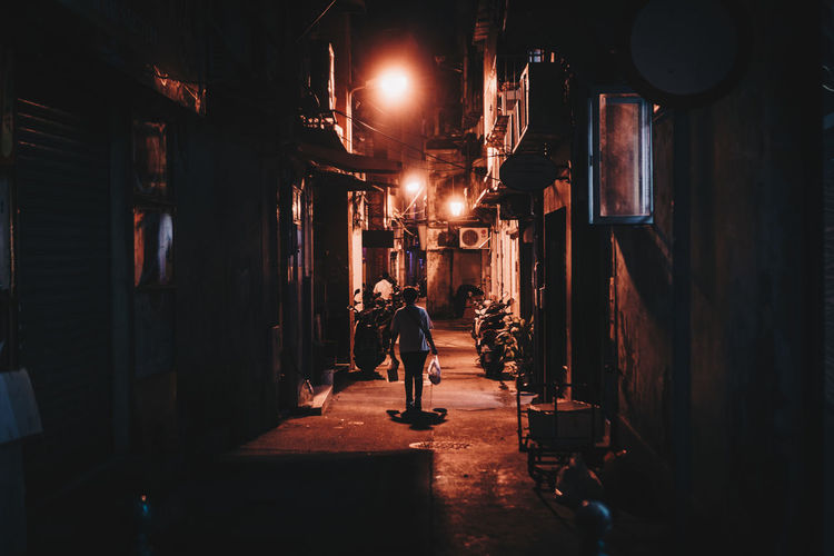 Rear view of woman walking on illuminated alley amidst buildings at night