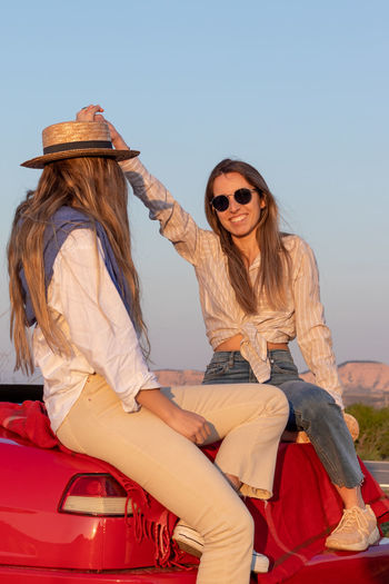 Vertical phoot of two friends enjoying spring break roadtrip sitting on a red car looking at sunset 