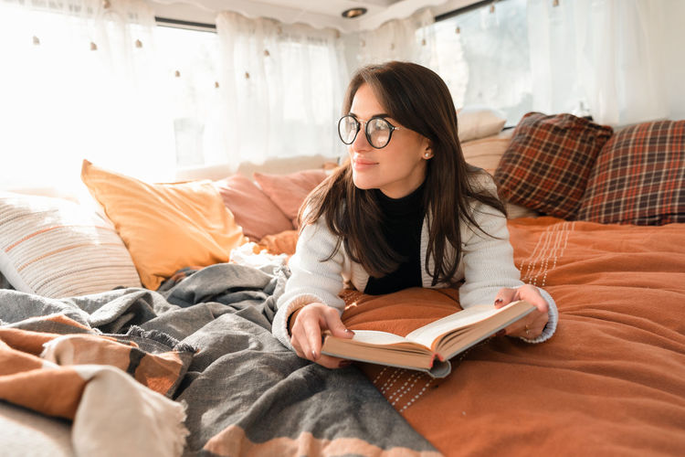 Brunette girl in glasses reading a book and lying on the bed in a camper van