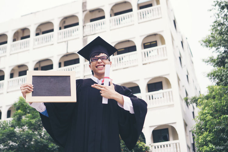 Smiling young man in graduation gown holding writing slate against building