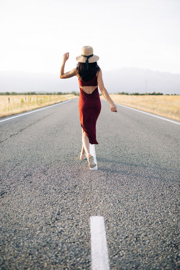 Back view traveler in summer dress and sunhat walking along empty roadway and looking away while enjoying freedom