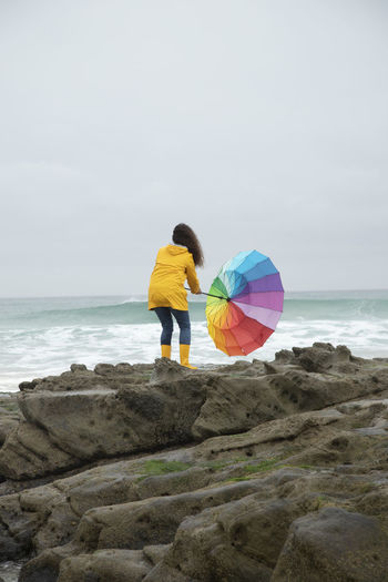 Woman holding umbrella in wind while standing on rock against sea