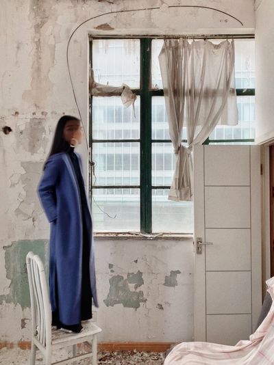 Young woman standing on chair while looking through window at abandoned home