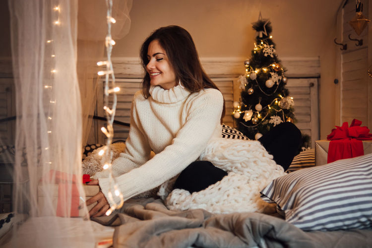Young woman sitting in illuminated christmas tree at home