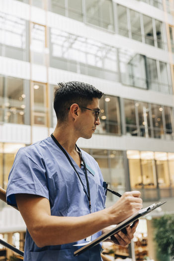Mature male healthcare worker looking away while writing in clipboard at hospital