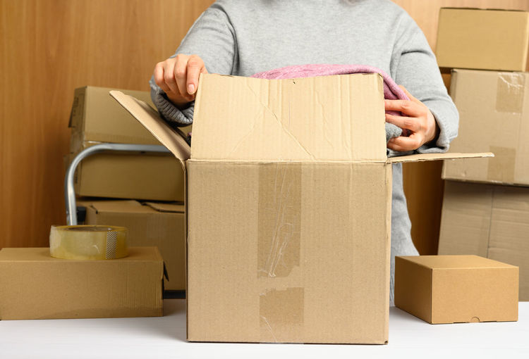 Woman in a gray sweater is packing brown cardboard boxes on a white table, behind a stack of box