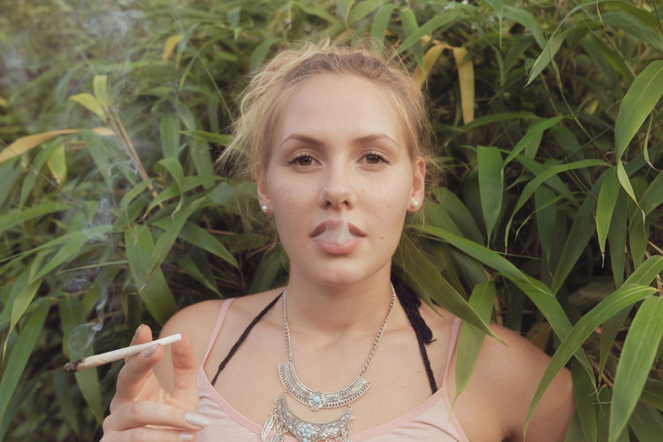 Portrait of young woman smoking marijuana joint while standing against plants