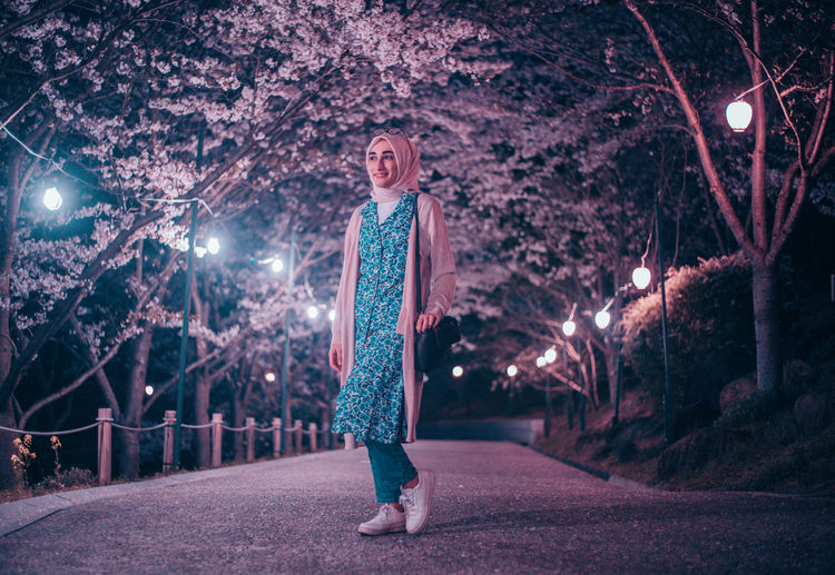 Portrait of young woman standing by illuminated trees at night