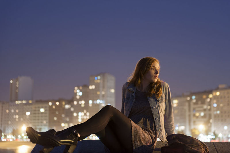 Young woman sitting on retaining wall against illuminated city at night