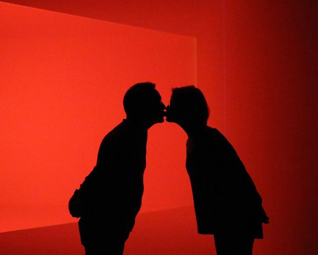 Side view of silhouette couple kissing on mouth while standing against red background