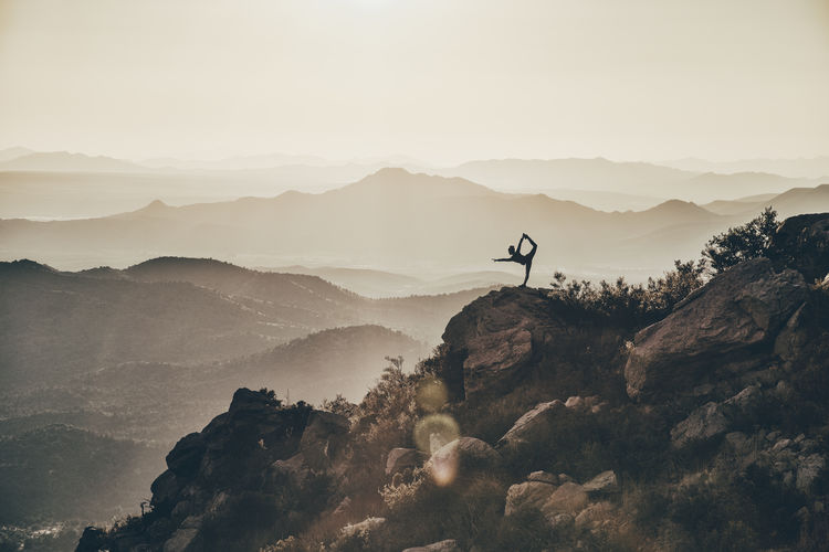 Mid distance view of silhouette woman exercising on rock against mountains during sunset
