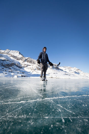 Young man playing ice hockey on frozen lake by snowcapped mountain against clear sky