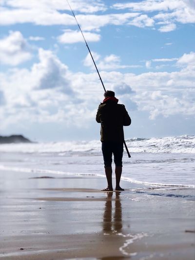 Rear view of man with fishing rod standing at beach against cloudy sky