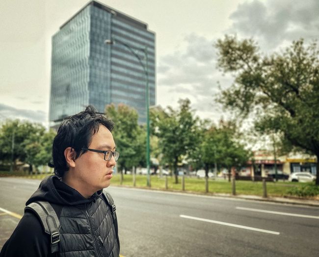 Side view of young asian man standing beside road against building in the city.