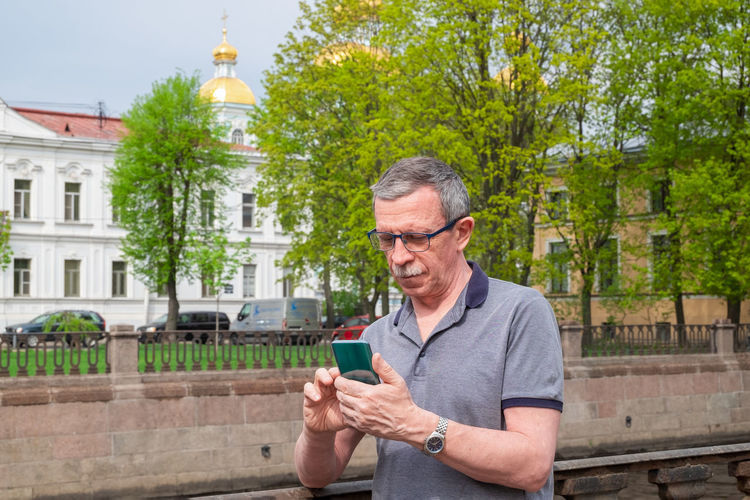 An adult caucasian senior man is typing a message on smartphone in the city 