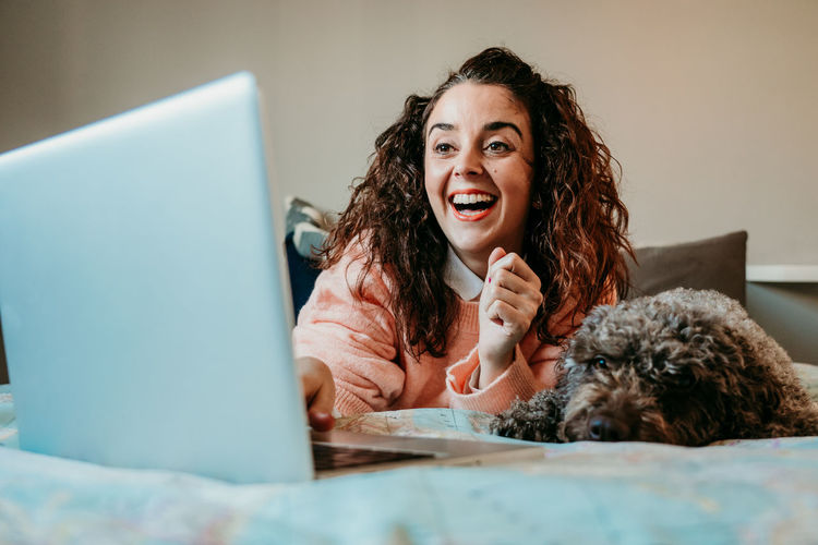 Smiling woman using laptop by dog on bed at home