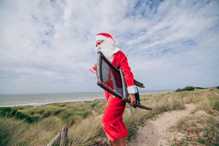 Santa claus walks on the dunes at the beach with a deck chair - on vacation before the christmas 