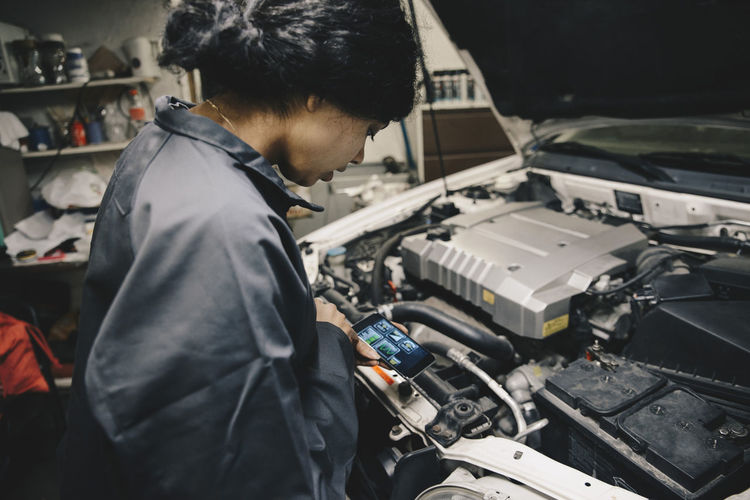 Female mechanic using application on smart phone while standing by open car hood