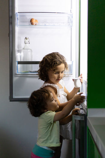 Little girl and toddler taking fresh yoghurt from fridge while stealing food at night at home together