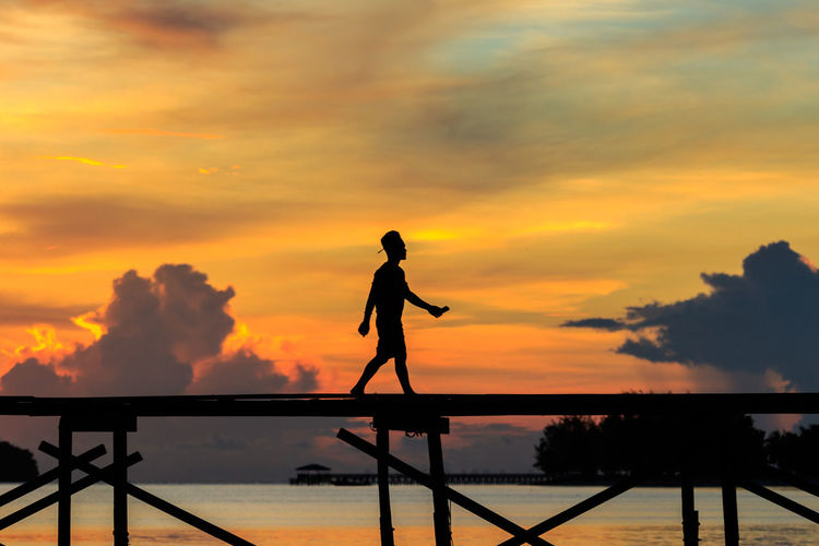 Silhouette man walking on bridge over sea against sky during sunset