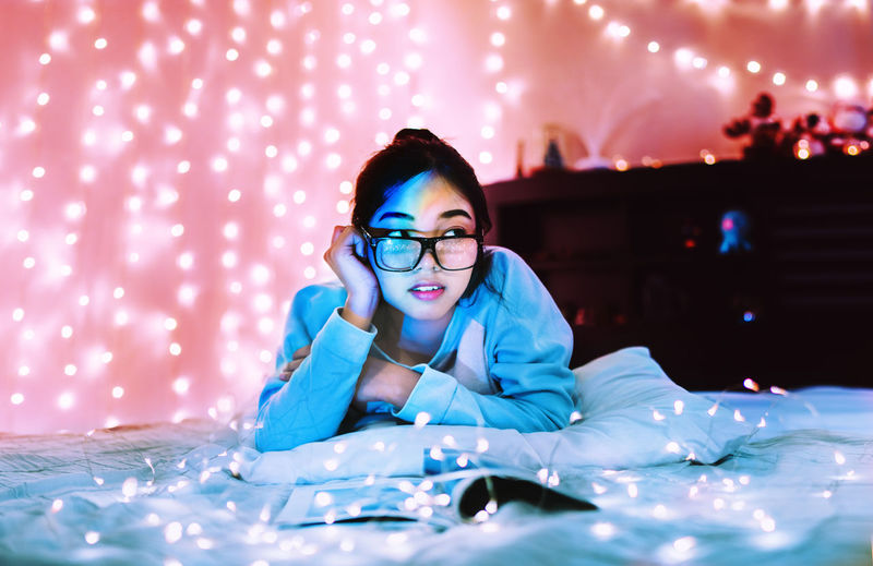 Young woman with illuminated string lights relaxing on bed during christmas